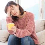 疲れた女性がソファに座り、頭を手で支えながらコーヒーカップを持っている様子。『疲れる』の英語表現を学ぶためのイメージ。