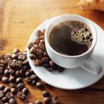 白いカップに注がれたブラックコーヒーと周囲に広がるコーヒー豆。クロワッサンとともに木製テーブルの上に置かれた、カフェタイムを楽しむシーン。