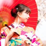 赤い和傘を差しながら桜の下でポーズを取る、華やかな浴衣姿の女性。浴衣の英語表現や日本文化を紹介する場面に最適。